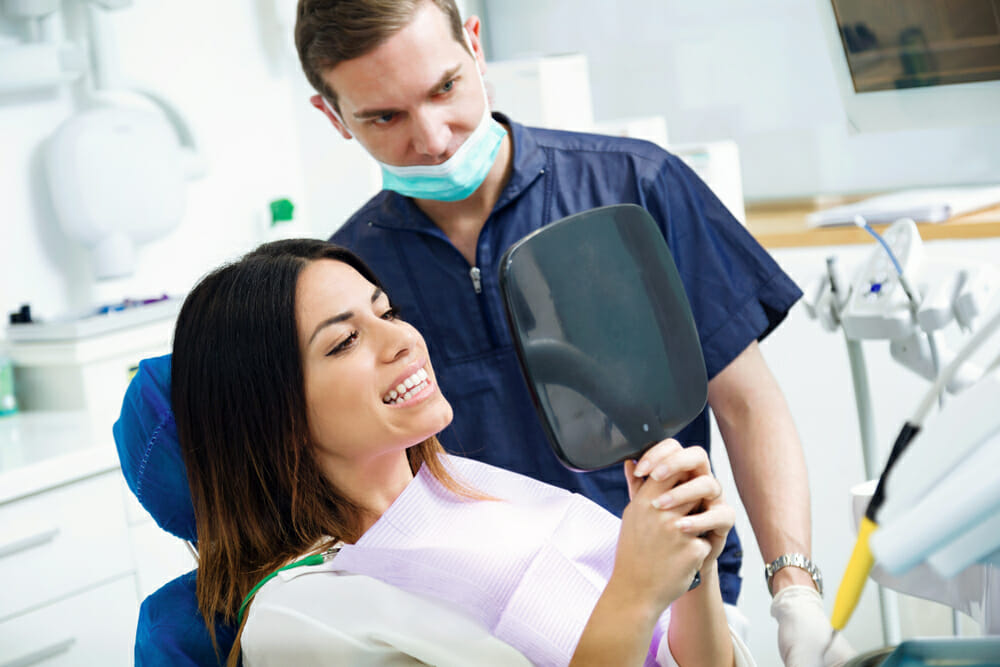 Woman Sitting in Dentist Chair Looking at Teeth in Handheld Mirror